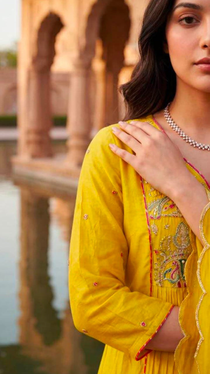 A woman wearing a bright yellow embroidered kurta with a matching skirt, standing in front of a blurred stone archway and water feature.